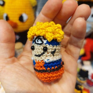 Peut inclure: Petite figurine au crochet faite à la main représentant un personnage avec des cheveux jaunes, un bandeau bleu et une tenue orange. La figurine a des yeux noirs et une expression souriante. La figurine est tenue dans une main.