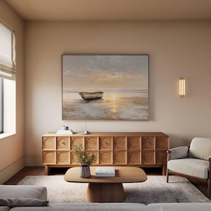 May include: A living room scene featuring a large framed painting of a boat on a beach at sunset. Below the painting is a wooden cabinet with a geometric design. A wooden coffee table and a gray armchair complete the room.