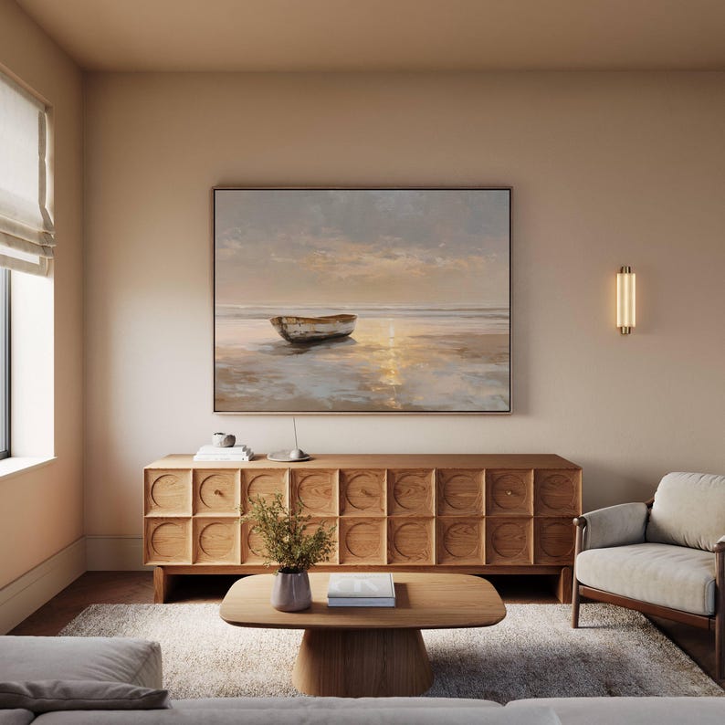 May include: A living room scene featuring a large framed painting of a boat on a beach at sunset. Below the painting is a wooden cabinet with a geometric design. A wooden coffee table and a gray armchair complete the room.