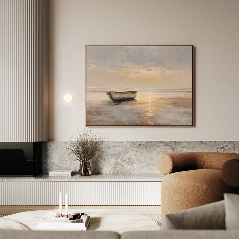 May include: A framed landscape painting depicts a small boat on a beach at sunset. The artwork features warm tones of gold, beige, and brown, with the boat's reflection visible in the water. The painting is displayed in a modern living room setting.