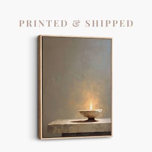 May include: A framed canvas print of a still life painting. The artwork shows a lit candle in a small bowl on a stone surface, against a muted, neutral background. The text "PRINTED & SHIPPED" is at the top.