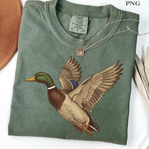 Op de afbeelding: Een saliegroen t-shirt met een gedetailleerde illustratie van een vliegende wilde eend. De eend heeft bruine, bruine, groene en blauwe veren. Een zilveren ketting met een kleine hanger rust op het shirt. Het shirt heeft een Comfort Colors-label.
