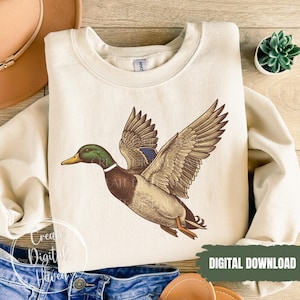 Op de afbeelding: Cr&egrave;mekleurige sweatshirt met een gedetailleerde illustratie van een vliegende wilde eend. De eend heeft bruine, groene en blauwe veren. De woorden "DIGITALE DOWNLOAD" zijn wit gedrukt op een groene rechthoek.