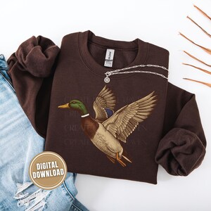Op de afbeelding: Een bruine sweatshirt met een gedetailleerde illustratie van een vliegende wilde eend. De eend heeft groene, bruine en blauwe veren. Een zilveren ketting met een hanger is over de sweatshirt gedrapeerd. Een "DIGITALE DOWNLOAD" sticker is zichtbaar.