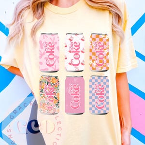Puede incluir: Una camiseta amarilla con seis latas de Diet Coke con varios diseños. Las latas son rosas, blancas y naranjas, con estampados florales, frutales y a cuadros. El logotipo de Diet Coke es visible en cada lata.
