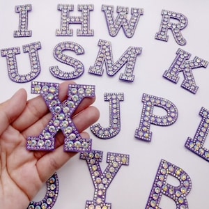 May include: A collection of purple, rhinestone-encrusted letters, with the letter "X" held in a hand. The letters are adorned with iridescent rhinestones, creating a sparkling effect. The letters are in various fonts and sizes, scattered on a white surface.