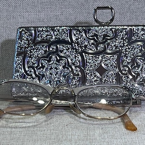 May include: A rectangular silver-toned metal clutch with an intricate embossed floral pattern and a top D-ring closure, paired with a pair of eyeglasses featuring silver-toned frames and translucent amber-colored temple tips.