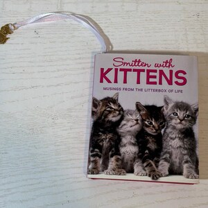 May include: A small book titled Smitten with KITTENS: Musings from the Litterbox of Life, featuring a photograph of four kittens on the cover. A white ribbon bookmark with a small gold-toned cat-shaped charm is attached to the top of the book.