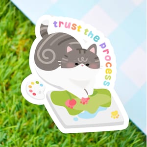 May include: A sticker featuring a cartoon cat painting on a canvas with a paint palette and the text "trust the process" in rainbow colors.