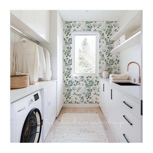 May include: A white laundry room with a white washing machine and tumble dryer, white cabinets, and a white worktop. The back wall is covered in a green and white floral wallpaper. There is a window in the back wall and a brown tap on the worktop.
