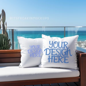 May include: Two white throw pillows with the text "YOUR DESIGN HERE" in blue on a white sofa with a wooden frame. The sofa is on a balcony overlooking a blue ocean.