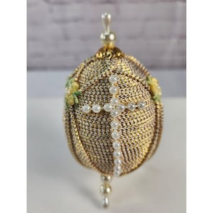 May include: An egg-shaped ornament covered in metallic gold and silver thread, featuring a cross made of small white faux pearls on the front. The ornament is topped and bottomed with a vertical arrangement of white faux pearls and gold trim.