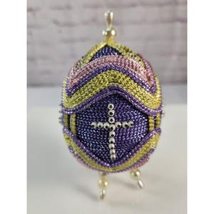 May include: An egg-shaped ornament featuring a purple, yellow, and pink woven fabric pattern. The center displays a cross made of small silver beads. The ornament is topped with a single pearl-like bead and stands on three small pearl-like feet.