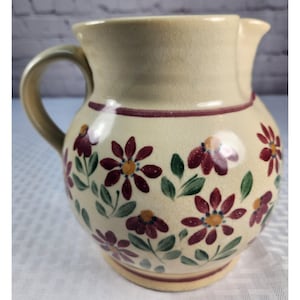 May include: A cream-colored ceramic pitcher featuring a hand-painted floral pattern with dark red petals, yellow centers, and green leaves. The pitcher has a rounded body, a curved handle, a flared rim, and a visible crackle glaze finish throughout.