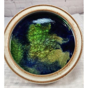 May include: Round ceramic dish with a cream-colored rim and a vibrant interior. The interior features a blend of green and blue hues, creating a textured, abstract design. The dish has a glossy finish.