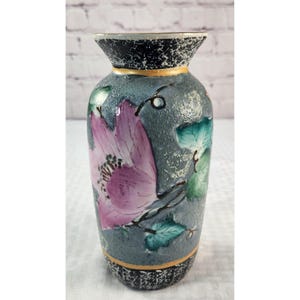 May include: A ceramic vase with a textured gray body featuring a hand-painted pink flower and green leaves. The rim and base have a black and white speckled pattern, and the neck and base are accented with thin gold-colored horizontal bands.