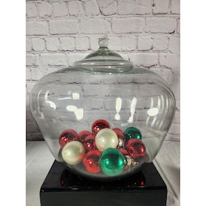 May include: A large, clear glass apothecary jar with a lid, filled with red, white, and green Christmas ornaments. The jar sits on a black base, with a white brick wall in the background. The ornaments are various sizes and have a shiny finish.