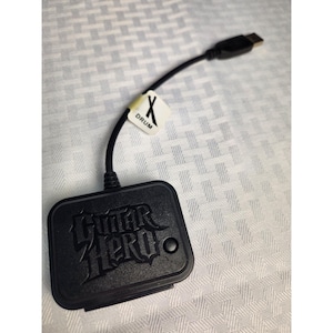 May include: Black Guitar Hero drum controller with a black cable and USB connector. The controller has the Guitar Hero logo embossed on the front. A white label with the word "DRUM" and an "X" is attached to the cable.