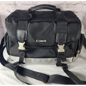 May include: Black Canon camera bag with a silver accent stripe and the Canon logo. The bag has a top handle, adjustable shoulder strap, and multiple compartments. Features metal buckles and leather accents.