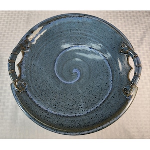 May include: A round ceramic serving platter with two integrated handles on opposite sides. The surface features a speckled blue glaze with a prominent lighter blue spiral pattern centered in the middle. The edges have a dark, mottled finish.