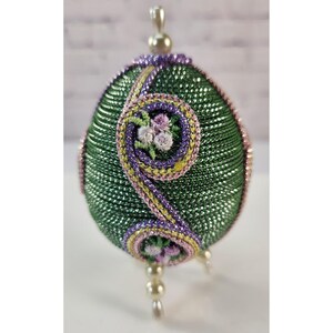 May include: An egg-shaped ornament wrapped in metallic green thread, featuring decorative swirls of purple and gold trim. The design includes small embroidered floral clusters in pink and white, accented by cream-colored pearl beads at the top and bottom.