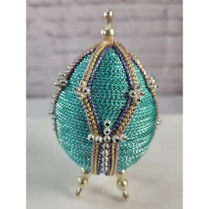 May include: A decorative egg ornament covered in shimmering teal metallic thread, featuring vertical bands of braided gold, pink, and purple trim. The egg is accented with small white faux pearls arranged in cross patterns and stands on three small pearl-tipped feet.