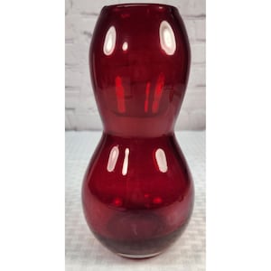 May include: A deep red glass vase with a smooth, glossy finish. The vase features a distinct hourglass shape with a wide, rounded base that tapers into a narrow middle section before widening again toward the open, circular top rim.