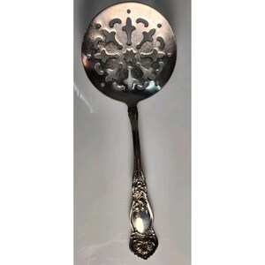May include: Silver-toned serving spoon with a decorative, pierced circular head. The head features a symmetrical, openwork design. The handle has an ornate floral pattern and a rounded, detailed end. A vintage kitchen utensil.