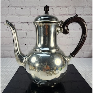May include: A polished silver-tone metal coffee pot featuring a rounded body, a long curved spout, and a dark brown handle with a matching finial on the lid. The surface is highly reflective, showing light highlights and a curved silhouette.