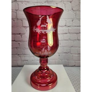 May include: A ruby red glass candle holder with a pedestal base. The glass features a frosted design of a winter scene with trees, a reindeer, and a building. The candle holder is approximately 8 inches tall.