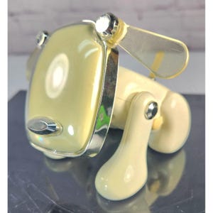 May include: A cream-colored plastic dog-shaped figurine featuring a smooth, rounded head with a chrome-finished border, a small oval chrome nose, and two translucent, pale yellow ear-like appendages attached to the sides with silver rivets.