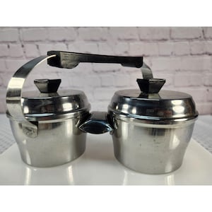 May include: A double-container condiment server made of polished stainless steel. It features two round bowls with matching lids, each topped with a black knob. A single black handle connects the two containers, allowing for easy transport.