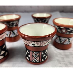 May include: A set of five ceramic shot glasses. The glasses have a white interior and a reddish-brown exterior with geometric patterns in black, white, and blue. The shot glasses are arranged on a white surface.