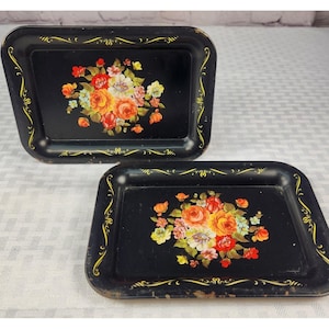 May include: Two rectangular black metal trays with floral designs. The trays feature a bouquet of orange, red, and yellow roses. The edges are decorated with gold scrollwork. The trays are likely vintage or antique.