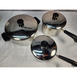 May include: A set of three stainless steel pots with lids. The pots have black handles and knobs. The lids are reflective, and the pots are arranged on a white surface.