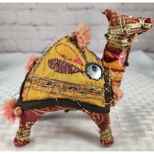 May include: A stuffed camel figurine crafted from patterned fabric in shades of yellow, red, and brown. The camel features gold metallic ribbon trim, small pink pom-poms, and a circular mirror accent on its side, with visible loose metallic threads.