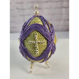 May include: A decorative egg ornament featuring a yellow metallic textured surface overlaid with purple braided cord patterns. A cross made of small white beads is centered on the front. The egg is topped with a pearl bead and stands on three pearl bead feet.