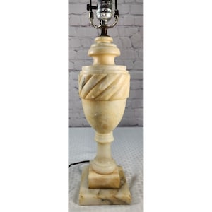 May include: A table lamp base crafted from cream-colored stone with a polished finish. The structure features a square base, a slender pedestal, a central urn-shaped body with diagonal fluted carvings, and a metal socket at the very top.