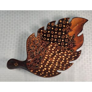 May include: A leaf-shaped wooden tray featuring a dark brown finish with intricate, hand-painted batik-style patterns. The design includes floral motifs, geometric shapes, and dotted details across the surface, with a smooth, curved edge.