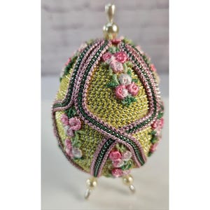 May include: An egg-shaped decorative ornament featuring a textured yellow metallic surface divided by pink and green braided trim. The surface is adorned with embroidered pink and white floral clusters, topped with a small pearl-like bead finial.