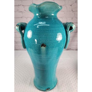 May include: A turquoise ceramic vase with a glossy crackle glaze finish, featuring a ruffled rim and two side handles. The body of the vase displays a distinct network of fine lines across its surface, with a small dark chip visible on the front.
