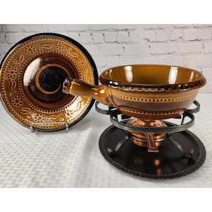 May include: A brown ceramic fondue pot with a handle, featuring a decorative embossed border, resting on a black metal stand with a burner. Beside it stands a matching brown ceramic plate with a dark center and an intricate floral pattern.