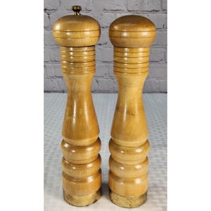 May include: Two tall, light-colored wooden pepper mills with a smooth, polished finish. Each mill features a rounded top, a series of horizontal grooves along the upper neck, and a wider, bulbous base. The bottom of each mill has a metallic band.
