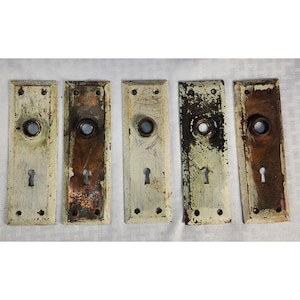 May include: Five rectangular metal door plates with a distressed, chipped off-white paint finish over a copper-toned base. Each plate features a central circular opening for a doorknob, a lower keyhole, and four small screw holes at the corners.
