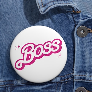 Może przedstawiać: Biały guzik z napisem "BOSS" w różowych literach z białym obramowaniem. Litery są w retro stylu czcionki i mają efekt 3D. Po obu stronach słowa znajdują się dwie małe białe gwiazdki.