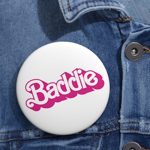 May include: A white button with a pink "Baddie" graphic. The button is pinned to a blue denim jacket.
