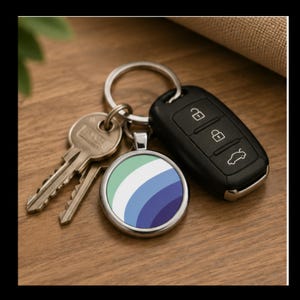 May include: A circular metal keychain featuring a gradient design of light green, white, light blue, and dark blue stripes. The keychain is attached to a silver split ring along with two standard silver-colored metal keys and a black plastic car key fob.