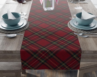 May include: A red plaid table runner with yellow and green accents, displayed on a wooden table set for a meal. The table is set with blue bowls, white plates, silverware, and wine glasses. A vase of pink flowers is in the center.