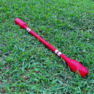 May include: A red and white wooden stick with a rounded end. The stick is lying on a bed of green grass.
