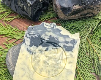 Puede incluir: Una barra de jabón artesanal con un diseño de remolino blanco y negro. El jabón está sobre un jabonera de piedra gris. El jabón tiene un logotipo circular que dice "Dream Soap" en blanco.